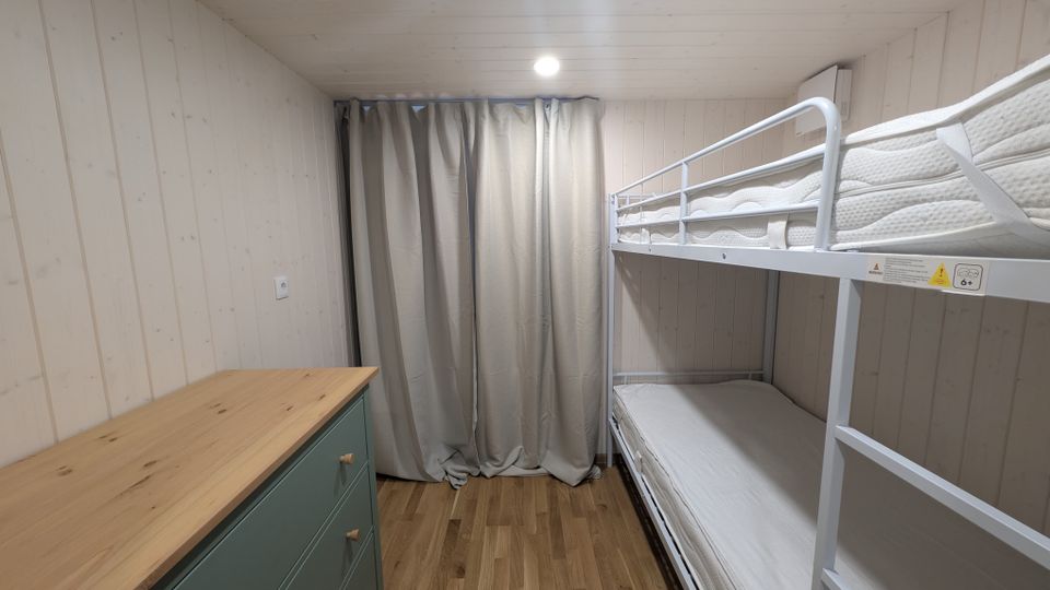 Kleines Schlafzimmer mit hellen Holzvertäfelungen an Wänden und Decke sowie Holzfußboden. Rechts steht ein weißes Metall-Etagenbett, links eine Kommode mit Holzplatte; am Ende des Raums hängt ein grauer Vorhang vor dem Fenster.