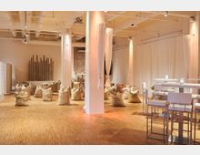 Ein heller Innenraum mit Holzboden, weißen Vorhängen und sichtbaren Deckenrohren. Der Raum enthält dekorative Säcke in der Mitte, hohe weiße Tische und Barhocker sowie schlichte Holz- und Backsteinelemente an den Wänden.