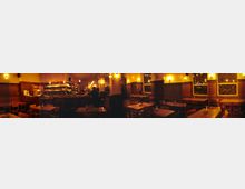 Ein gemütliches Restaurant oder Café mit Holzmöbeln, warmem gelbem Licht und dekorativen Wandleuchten. Im Hintergrund ist eine Bar mit Regalen voller Gläser und Flaschen sowie mehrere verglaste Fenster mit Gittern zu sehen.