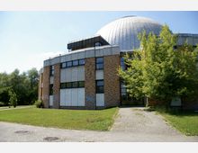 Gebäude mit einer Kuppel und Fassade aus Backstein und Beton, umgeben von einer Grünfläche mit Bäumen; gepflasterter Weg führt zum Eingangsbereich.