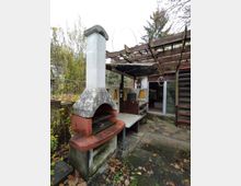 Außenbereich mit gemauertem Grill aus Beton und Ziegeln, überdacht von einer hölzernen Pergola. Im Hintergrund sind eine verglaste Tür, ein Treppenaufgang und teilweise bepflanzte Flächen sichtbar, bedeckt von fallendem Herbstlaub.