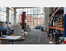 Industriehalle mit großer Fensterfront, durch die ein Backsteingebäude und Bäume sichtbar sind. Im Innenraum sind Arbeitsstationen, Maschinen, Regale mit Paletten und Materialien sowie ein Schreibtisch mit Monitor zu sehen; im Hintergrund steht ein schwarzes Auto.