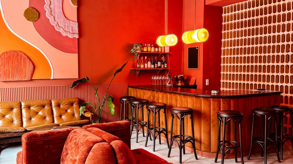 Modern lounge bar interior with red walls, a curved rust-colored sectional sofa, and a wooden bar lined with tall black stools under hanging globe pendant lights. The space includes an abstract wall painting, a small back bar shelf, and a patterned brick screen along one side.