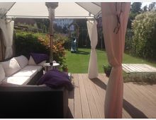 Sonniger Gartenbereich mit überdachter Terrasse, ausgestattet mit einer weißen Sitzgarnitur und einem Heizstrahler. Im Hintergrund ist eine Rasenfläche mit einer Schaukel und einer Rutsche zu sehen, umgeben von Hecken und Pflanzen.