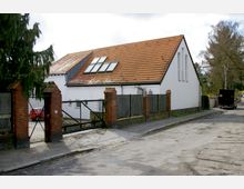 Ein einstöckiges Gebäude mit weißer Fassade und rotem Ziegeldach, ausgestattet mit großen Dachfenstern, hinter einem Zaun mit Backsteinpfeilern und Metalltor. Rechts im Bild verläuft eine asphaltierte Straße mit parkenden Fahrzeugen, umgeben von einigen Bäumen.