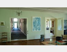 Innenansicht eines Gebäudes mit hellgrünen Wänden und Holzboden. Links führt ein schmaler, dunkler Flur mit Beleuchtung zu einer Tür, rechts sind zwei Glastüren geöffnet und zeigen einen Wohnraum mit Büchern und Möbeln.