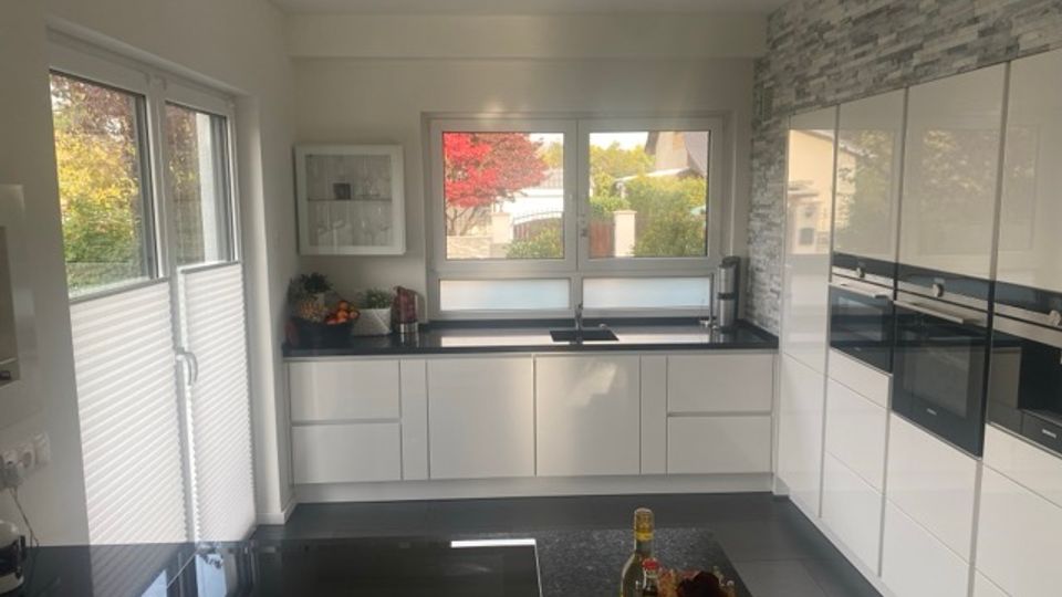 Moderne, helle Küche mit weißen, griffleistenlosen Schränken, schwarzen Arbeitsflächen und integrierten Backöfen an einer Wand mit grauer Steinoptik. Große Fenster und eine Glastür lassen viel Tageslicht herein; im Vordergrund steht eine Kücheninsel mit schwarzer Platte.