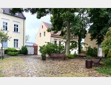 Kleiner gepflasterter Platz mit Holzbänken und Begrünung, umgeben von gelblichen Wohngebäuden mit roten Ziegeldächern. Große Bäume spenden Schatten, und ein Mülleimer steht in der Nähe der Bänke.