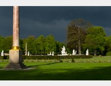 Ein gepflegter Barockgarten mit einer Marmorsäule im Vordergrund, umgeben von Statuen und Hecken, vor einer Kulisse aus grünen Bäumen und dramatischem, dunklem Himmel.