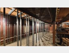 Innenansicht eines Baustellenbereichs mit Holz- und Stahlträgern. Der Raum hat unbehandelte Holzböden und Wände, sowie freigelegte Metallstreben, die die Struktur des Gebäudes stützen.