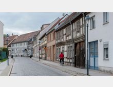 Pflasterstraße in einer ruhigen Altstadt mit Fachwerkhäusern, teils renoviert, teils mit Holzstrukturen. Eine Person fährt mit einem Fahrrad am Gehweg, umgeben von Straßenlaternen und Häusern mit roten Ziegeldächern.