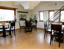 Heller Innenraum eines Cafés oder Restaurants mit Holzboden, mehreren Tischen und dunkelbraunen Stühlen. Große Fenster mit Holzrahmen lassen Tageslicht herein, an den Wänden hängen schwarz-weiße Bilder, und eine Treppe führt in ein Obergeschoss.