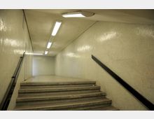 Treppe in einem schlichten U-Bahn- oder Fußgängertunnel mit beige gefliesten Wänden, einem schwarzen Handlauf und rechteckigen Deckenleuchten. Der Tunnel endet in einem ebenen Bereich mit heller Beleuchtung.