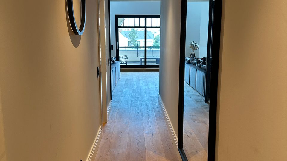 Langer, schmaler Flur in einer modernen Wohnung mit hellem Holzboden, beigen Wänden und Decken-Spotlights. Rechts hängt ein großer Spiegel, links eine runde Wandspiegel-Dekoration; am Ende führen Glastüren zu einem Wohnbereich mit Blick nach draußen.