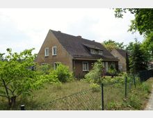 Zweistöckiges Wohnhaus mit brauner Putzfassade und dunklem Satteldach, umgeben von einem Garten mit Bäumen und Büschen. Im Vordergrund ein Maschendrahtzaun, im Hintergrund weitere ähnlich gestaltete Häuser.