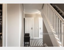 Heller Hausflur mit einer weißen Treppe, die nach oben führt, schwarz-weiß kariertem Fliesenboden und zwei weißen Türen mit klassischen Türgriffen. Links steht ein schwarzer Polsterstuhl an der Wand.