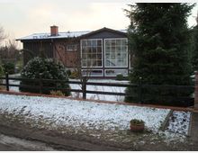 Einstöckiges Wohnhaus mit einer Backsteinfassade und großen Fenstern, umgeben von einem Garten mit Schneebedeckung. Im Vordergrund sind ein Holzzaun, Pflanzen und ein Tannenbaum zu sehen.