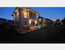 Beleuchteter Garten hinter einem modernen zweigeschossigen Einfamilienhaus in der Dämmerung, mit Holzterrasse, Esstisch und Stühlen sowie Einbauspots im Deck und Fassadenleuchten. Rasenfläche mit Sträuchern und Gartenbeleuchtung, im Hintergrund weitere Wohnhäuser.