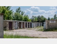 Eine Reihe von schlichten Garagen mit Flachdächern aus Beton, die aneinander gereiht sind, umgeben von Bäumen und Grünflächen. Ein Schild mit der Aufschrift „Garagenmarkt“ ist an der rechten Wand angebracht, und der Boden davor ist unbefestigt.