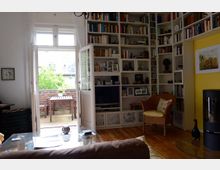 Wohnzimmer mit einer hohen Bücherwand, einem Fernseher und einem Kamin. Der Raum hat einen Holzfußboden, helle Wände und führt durch eine geöffnete Glastür auf einen Balkon mit Tisch und Stühlen, umgeben von Pflanzen.