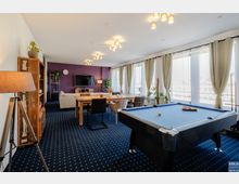 Wohnraum mit modernen Möbeln, einer lila Wand mit Fernseher, einem großen Esstisch und einem Billardtisch im Vordergrund. Große Fenster mit beigen Vorhängen lassen viel Tageslicht in den Raum, der mit blauem Teppichboden ausgestattet ist.