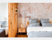 Modernes Schlafzimmer mit einer natürlichen Holztrennwand, die den Raum in zwei Bereiche teilt. Rechts steht ein Bett mit blauer Bettwäsche vor einer Wand mit floraler Tapete, daneben ein Nachttisch mit kleiner Lampe.