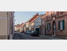 Schmale Kopfsteinpflasterstraße mit historischen Häusern auf beiden Seiten, darunter Gebäude aus roten Ziegeln und Fachwerk. Autos sind entlang der Straße geparkt, und ein Straßenschild mit der Aufschrift „Breite Straße“ ist sichtbar.