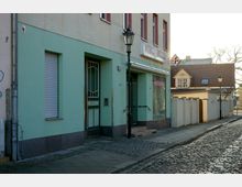 Fassadenansicht eines zweistöckigen Gebäudes mit mintgrüner Unterfassade und beigefarbenem Oberstock. Links eine Eingangstür, rechts ein Schaufenster, ein straßenbeleuchtender Laternenpfahl, gepflasterter Bürgersteig und eine Kopfsteinpflasterstraße. Im Hintergrund weitere Häuser mit roten Dächern.