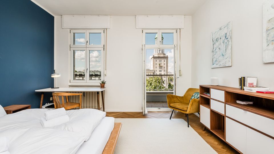Helles, modern eingerichtetes Schlafzimmer mit bodennahem Bett, Schreibtisch am Fenster und Sideboard an der rechten Wand. Zwei hohe Fenster und eine Balkontür führen nach draußen; Parkettboden im Fischgrätmuster, blauer Akzentwandbereich und Blick auf ein markantes Gebäude in der Ferne.