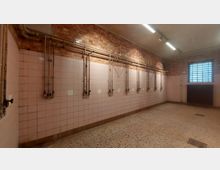 Ein leerer, funktionaler Duschraum mit hellen Wandfliesen, freiliegenden Rohrleitungen und mehreren Duscharmaturen an einer Ziegelwand. Der Boden ist mit kleinen, grauen Mosaikfliesen bedeckt, und ein vergittertes Fenster sowie eine Dunkelheizung sind sichtbar.