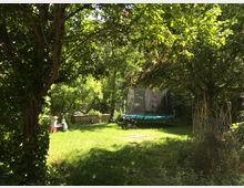 Grüner Garten mit großer Rasenfläche unter dichtem Blätterdach, umgeben von Bäumen und Sträuchern. Im Hintergrund stehen ein rundes Trampolin mit Sicherheitsnetz sowie eine niedrige Holzbegrenzung am Rand des Grundstücks.
