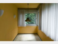 Kleiner Raum mit gelb gestrichenen Wänden, bodentiefen Fenstern mit weißen Vorhängen und einer hängenden Deckenleuchte mit glasartigem Lampenschirm. Durch die Fenster ist dichter, grüner Baumbewuchs zu sehen.