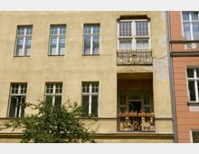 Fassade eines mehrstöckigen Altbaus mit gelber Putzwand, mehreren rechteckigen Fensterreihen und einem kleinen Balkon mit verzierten Metallgeländern. Ein Baum mit grünen Ästen ragt vor dem Gebäude hervor, und rechts ist ein angrenzendes, rötliches Nachbargebäude sichtbar.