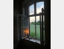 Ein offenes Holzfenster mit abgeblättertem Lack zeigt eine Aussicht auf eine grüne Wiese mit Bäumen im Hintergrund bei Sonnenuntergang. Auf der Fensterbank stehen eine brennende Kerze in einer Flasche, ein Glas und ein Buch.