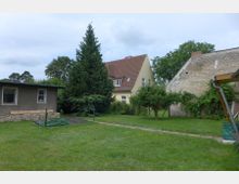 Grasbewachsener Hofgarten mit schmalem Weg, hohen Nadel- und Laubbäumen sowie einigen Obstbäumen. Im Hintergrund stehen ein zweigeschossiges Wohnhaus mit rotem Satteldach, ein flacher Anbau links und eine große gemauerte Scheunenwand rechts, teils mit Kletterpflanzen bewachsen.