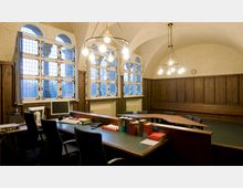 Büro- oder Besprechungsraum mit gewölbter Decke und großen Fenstern mit Gitterdesign. Holzwände, eine lange Bank, ein zentraler Tisch mit Stühlen und eine Arbeitsstation mit Büchern und Computer prägen den Raum.