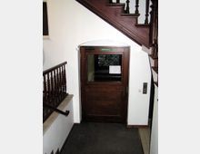 Holztür mit einem kleinen Fenster und einem grünen Schild mit der Aufschrift „Notausgang“ in einem Treppenhaus mit dunklem Holzgeländer. Die Szene zeigt weiße Wände, Teppichboden und einen Aufzugsknopf an der Wand rechts der Tür.