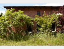 Backsteingebäude mit flachem Dach, teilweise von dichtem Grün und blühenden Sträuchern umgeben, im Vordergrund hohe Gräser und wilde Vegetation.