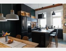 Moderne Küche mit dunklen Einbaumöbeln, einer freistehenden Kochinsel und hölzernen Akzenten. Im Vordergrund steht ein Esstisch mit Blumen und Obstschale, während Licht durch drei Fenster mit weißen Vorhängen einfällt.