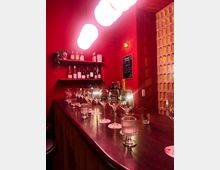 Small bar interior with deep red walls, a long dark wooden counter set with wine glasses and candle holders, and shelves of wine bottles and spirits behind the bar. Round pendant lights hang overhead, and a decorative block-pattern partition lines one side.
