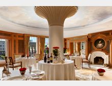 Eleganter Speiseraum mit rund angeordneter Tischdekoration, einem zentralen Pfeiler mit fächerförmigem Dekor an der Decke und großen Fenstern, die den Blick auf das Brandenburger Tor bieten. Die klassische Einrichtung umfasst Holzvertäfelungen, goldene Vorhänge, einen dekorativen Kamin sowie Tischdekorationen mit roten Rosen.