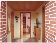 Flur in einem Wohnhaus mit holzgetäfelter Decke und Fliesenboden; an der rechten Wand ein kleines Hängeregal mit Pflanze, links ein Spiegel. Im Hintergrund ein geöffneter Durchgang zu einem weiteren Zimmer mit Holzmöbeln und einem roten Stuhl. Die linke Wand ist teilweise aus rotem Backstein.