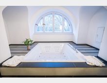 Helles Wellness-Badezimmer mit einem eingelassenen Whirlpool in gefliester Umrandung und integrierten Düsen. Dahinter befindet sich ein großes Rundbogenfenster, umgeben von weiß verputzten, gewölbten Wänden.