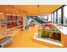 Modern gestalteter Innenraum mit orangefarbenem Boden und Decke, großen Fensterfronten und einer Glastrennwand mit gepunktetem Muster. Im Raum befinden sich ein Aquarium mit Pflanzen, Sitzgelegenheiten sowie eine Wand mit einem großflächigen Bild des Weltalls, das Planeten und Sterne zeigt.