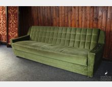 Grünes, gepolstertes Sofa mit Knopfdetail vor einer Holzwand. Im Hintergrund ist ein roter Paravent mit goldenen Mustern zu sehen, der Raum hat einen grauen Teppichboden.