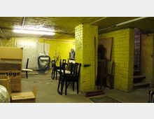 Ein gelb gestrichener Kellerraum mit sichtbaren Ziegelwänden, einer offenen Deckenbeleuchtung und vereinzelten Möbeln wie Stühle und ein Tisch. Im Raum befinden sich zudem Kartons, ein Besen und eine Waschbeckeninstallation an der Rückwand; rechts führen schmale Treppen hinauf.