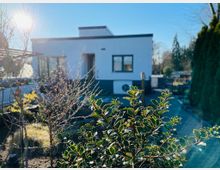 Ein modernes, einstöckiges Gebäude mit weißer Fassade und flachem Dach, im Vordergrund ein Vorgarten mit niedrigen Büschen und kahlen Sträuchern. Im Hintergrund sind Bäume und klare Himmel zu sehen, das Sonnenlicht fällt von links in die Szene.