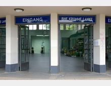 Eingangsbereich des U-Bahnhofs "Krumme Lanke" mit offenen Glastüren, grün gefliesten Wänden, einer Uhr und Rolltreppen im Hintergrund. Rechts im Bild befindet sich ein kleiner Blumenladen.