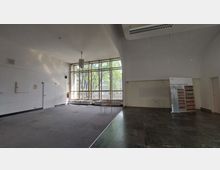 Großer, leerer Innenraum mit hohen Decken, teilweise Teppich- und Fliesenboden sowie einer großen Fensterfront mit gelbem Rahmen, die den Blick auf Bäume und ein Gebäude im Hintergrund freigibt. Ein einzelner Stuhl steht vor der Fensterfront, rechts ein Rollbanner mit der Aufschrift „Stasi-Unterlagen-Archiv“.