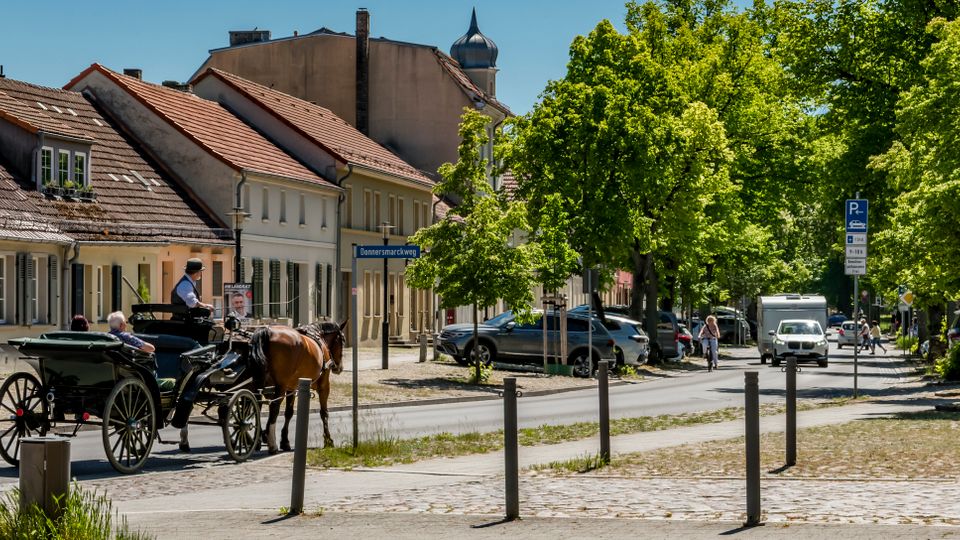 Straßenszene in einer Kleinstadt mit niedrigen, pastellfarbenen Häusern und roten Ziegeldächern entlang der Fahrbahn. Rechts säumen große, dicht belaubte Bäume den Straßenrand mit geparkten Autos, Pollern und einem Gehweg; im Hintergrund ragt ein Kirchturm über die Dächer.